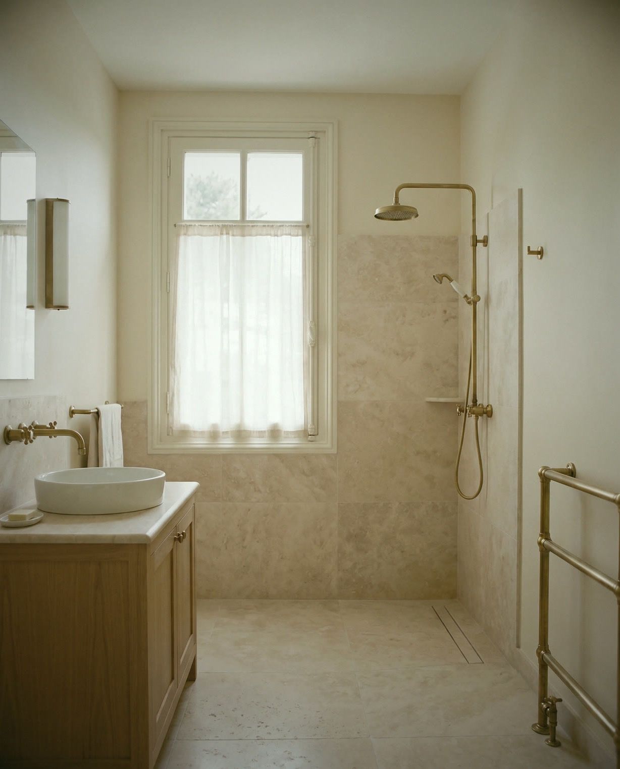 Salle de bain en travertin clair : vasque sur meuble de chêne, robinetterie et douche en laiton, fenêtre voilée.