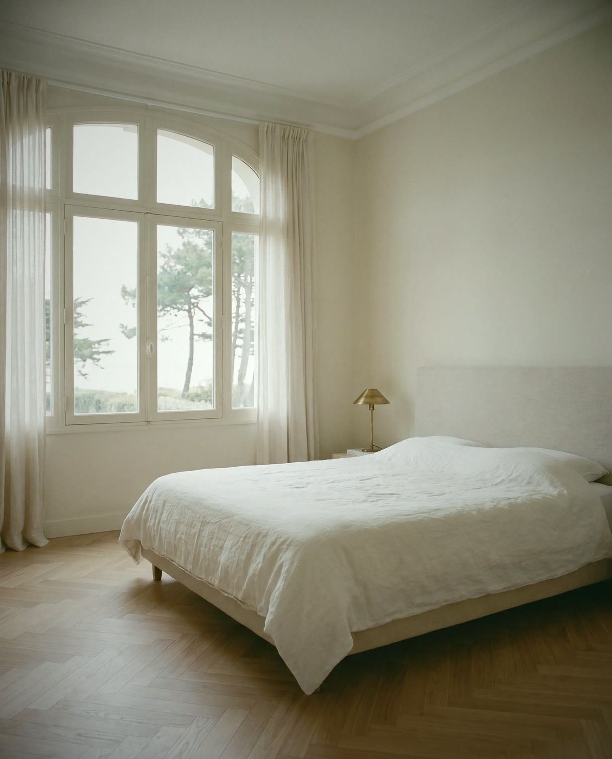 Chambre claire : tête de lit en lin, parquet chevron, haute fenêtre cintrée sur les pins maritimes.