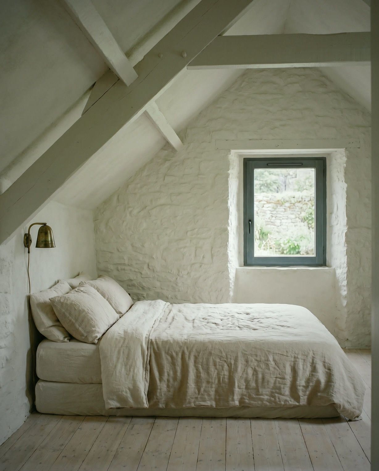 Chambre sous charpente, murs de pierre chaulée, lit en lin, applique en laiton, fenêtre ardoise sur le muret de pierre.