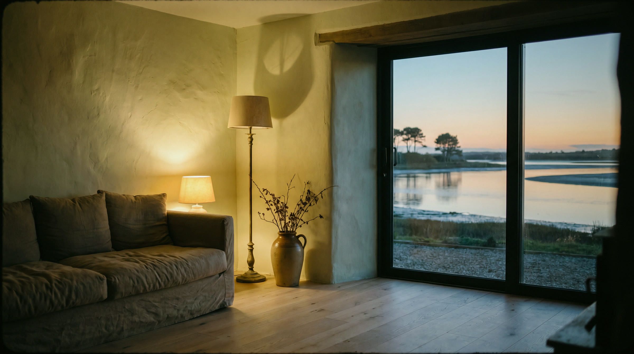 Intérieur d’une maison de l’Atelier au crépuscule : lumière chaude, grande baie sur l’estuaire du golfe du Morbihan, mur de plâtre clair et canapé de lin.