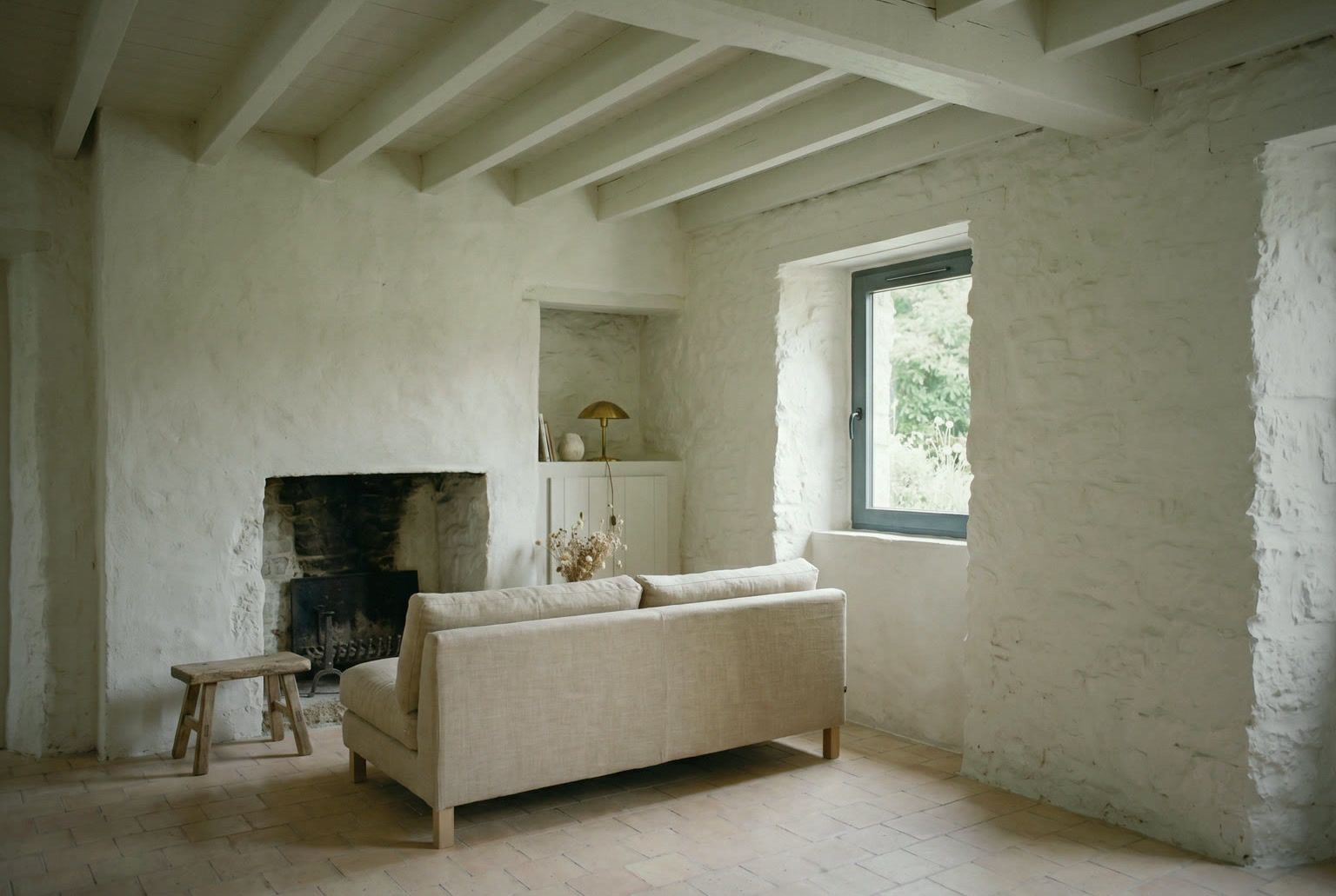 Pièce de vie du penty vue d’ensemble : cheminée, mur de pierre chaulée, canapé en lin, tabouret de bois et poutres peintes en blanc.